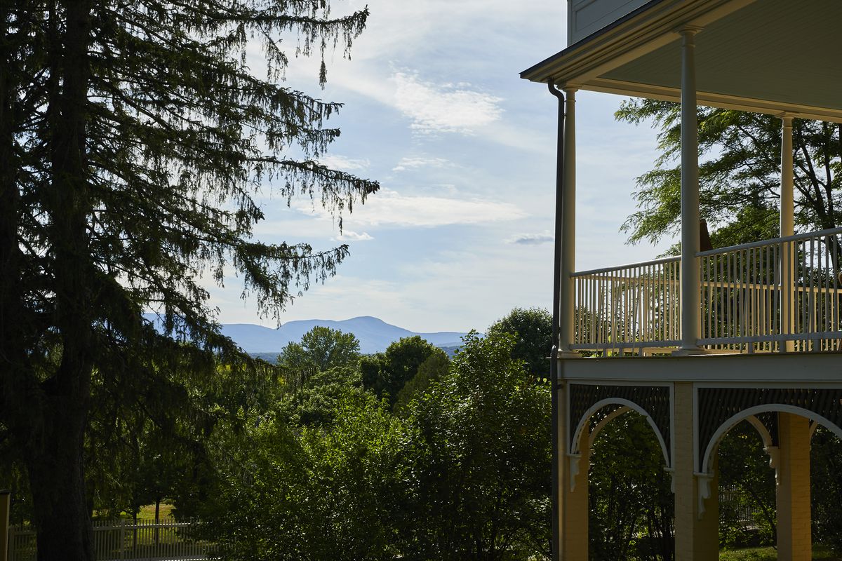 There is a house with an outside porch in the foreground. In the distance are trees and mountains.