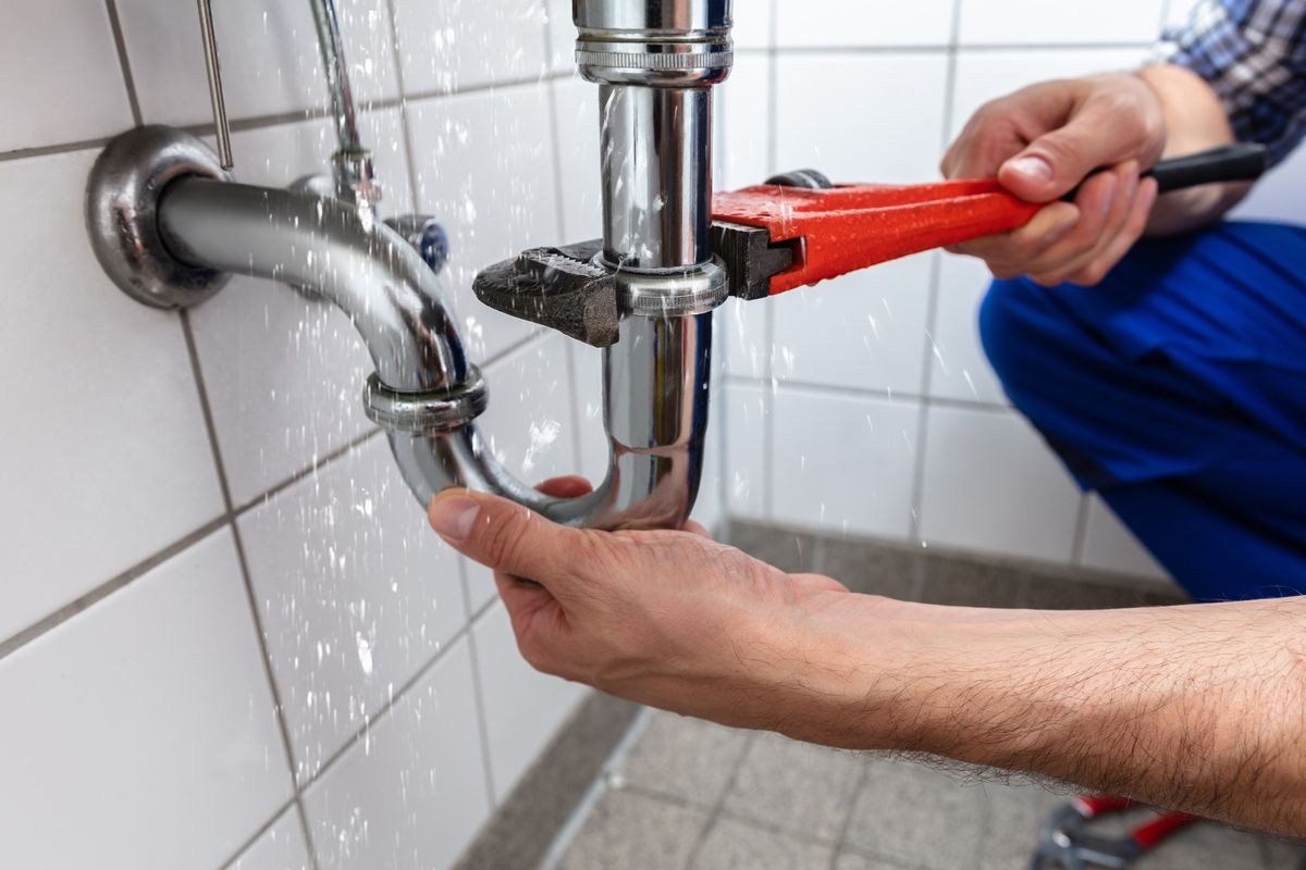 A plumber wearing blue pants uses a red wrench to fix a silver pipe beneath a sink in a white tile bathroom. 