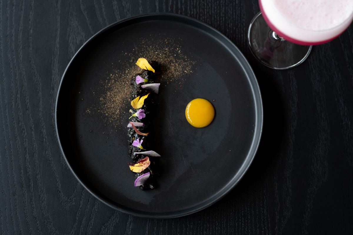 A stark black dish with a black central line of charred bits decorated with pops of yellow and lavender. A pool of golden sauce sits off to the side and a faint dusting of gold powder decorates the top left side of the plate.