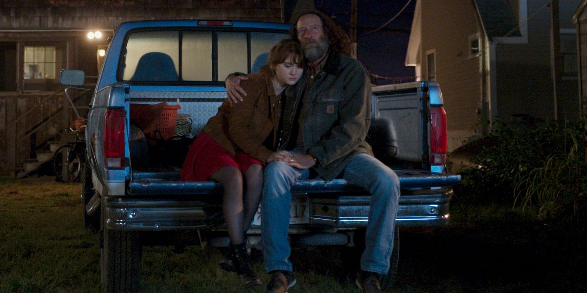 A teenaged girl and her father sit in the back of a truck. He has his arm around her shoulders.