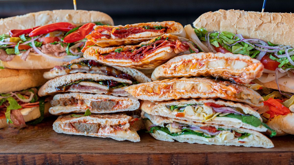 Italian paninis and subs are stacked on top of each other, all coated in sauces and filled with various, colorful meats and vegetables.