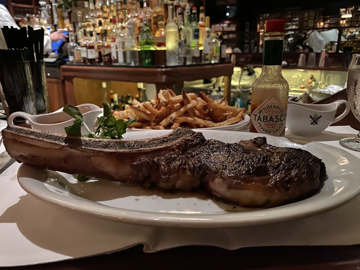 A bone-in rib steak on a white plate with fries and Tabasco sauce in the background.