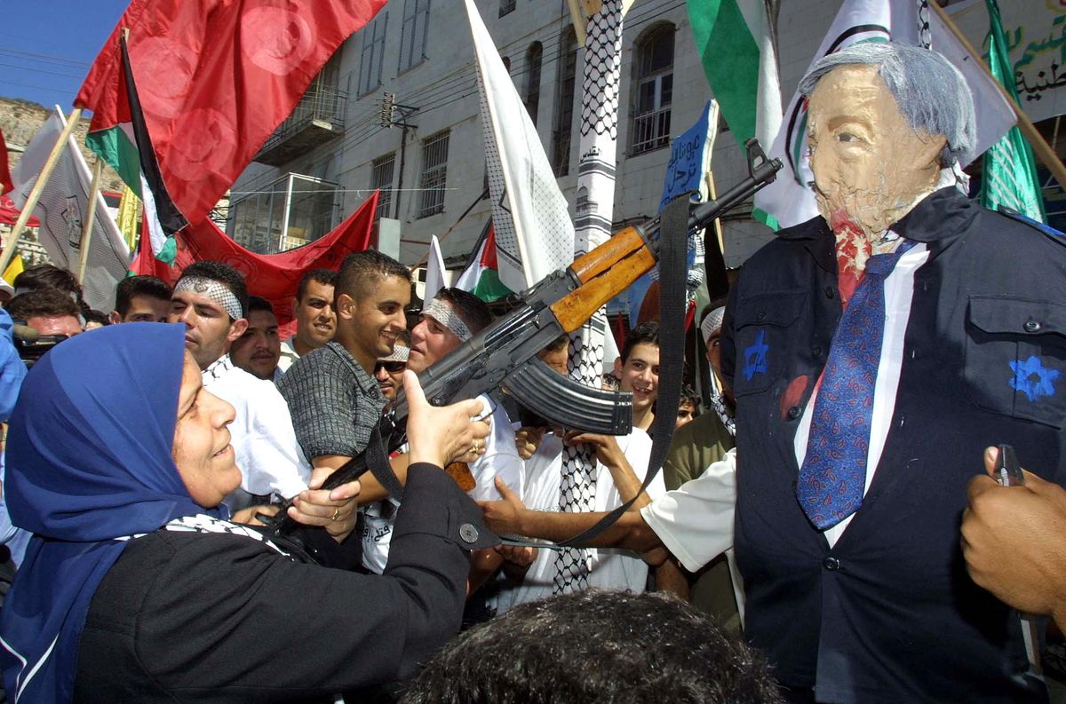 The woman holding the rifle smiles, its muzzle pointed at the paper mache face of Sharon, who is dressed in a black jacket featuring painted-on blue stars of David and red blood. A crowd parties behind her, many holding Palestinian flags.