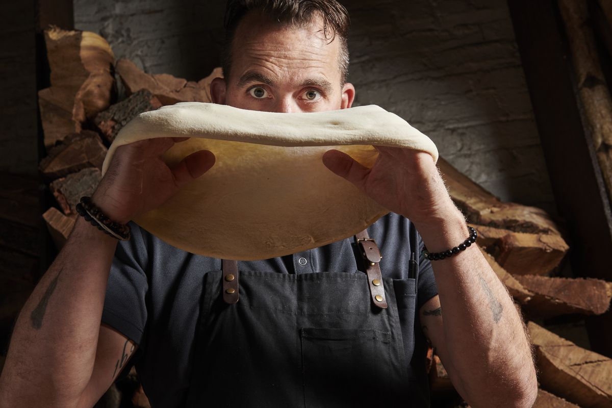 A man stands holding pizza dough up to eye level and staring at the camera.