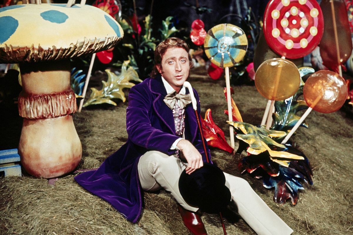 Wilder dressed in a purple coat as Wonka sits among oversize lollipops and mushrooms.