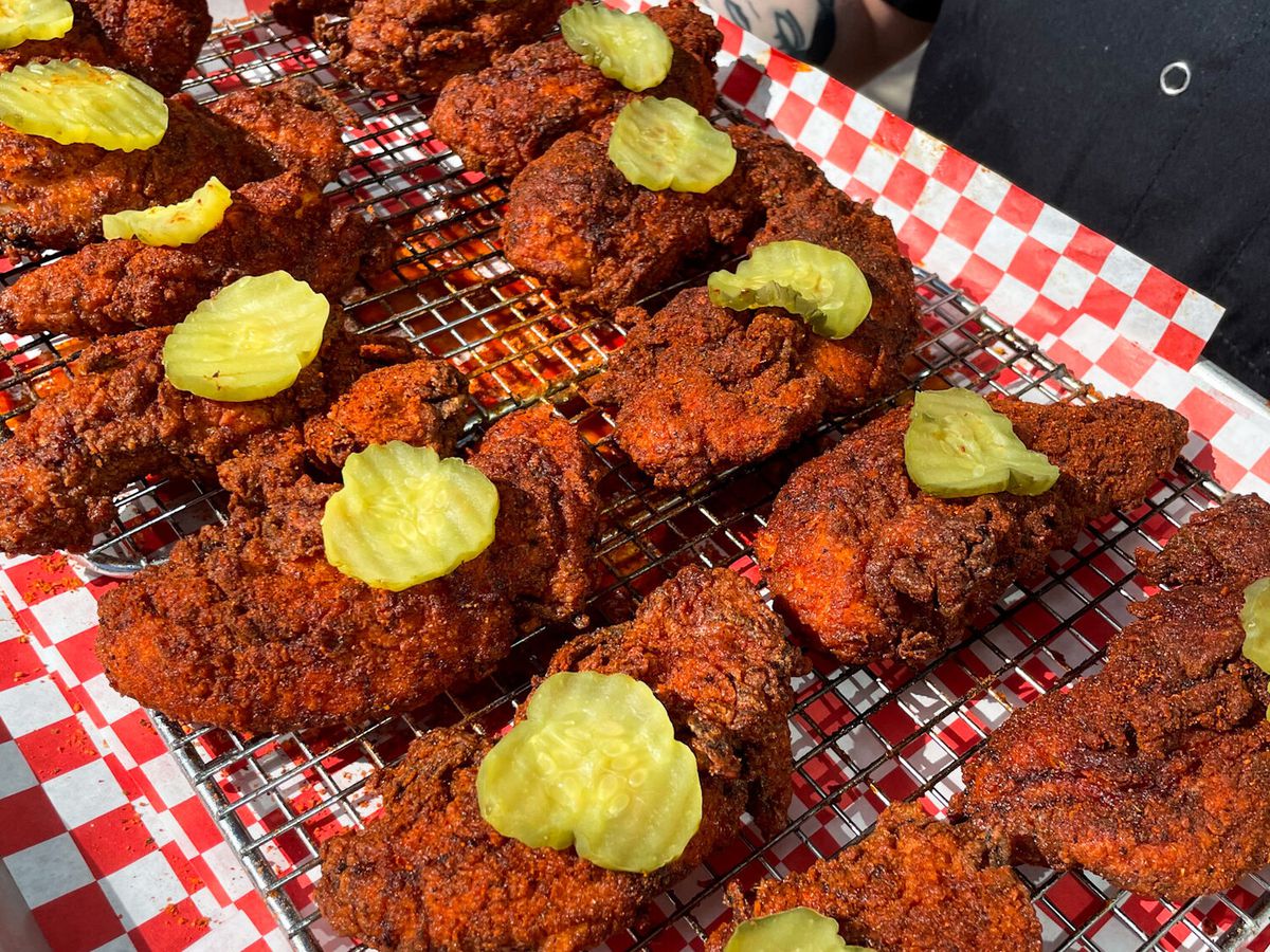 Fried chicken filets on a metal grate above red and white-checkered paper, all topped with a single pickle slice.