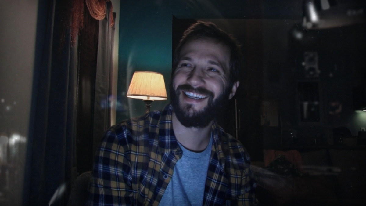 A man with a beard in a bedroom looks off-camera and laughs