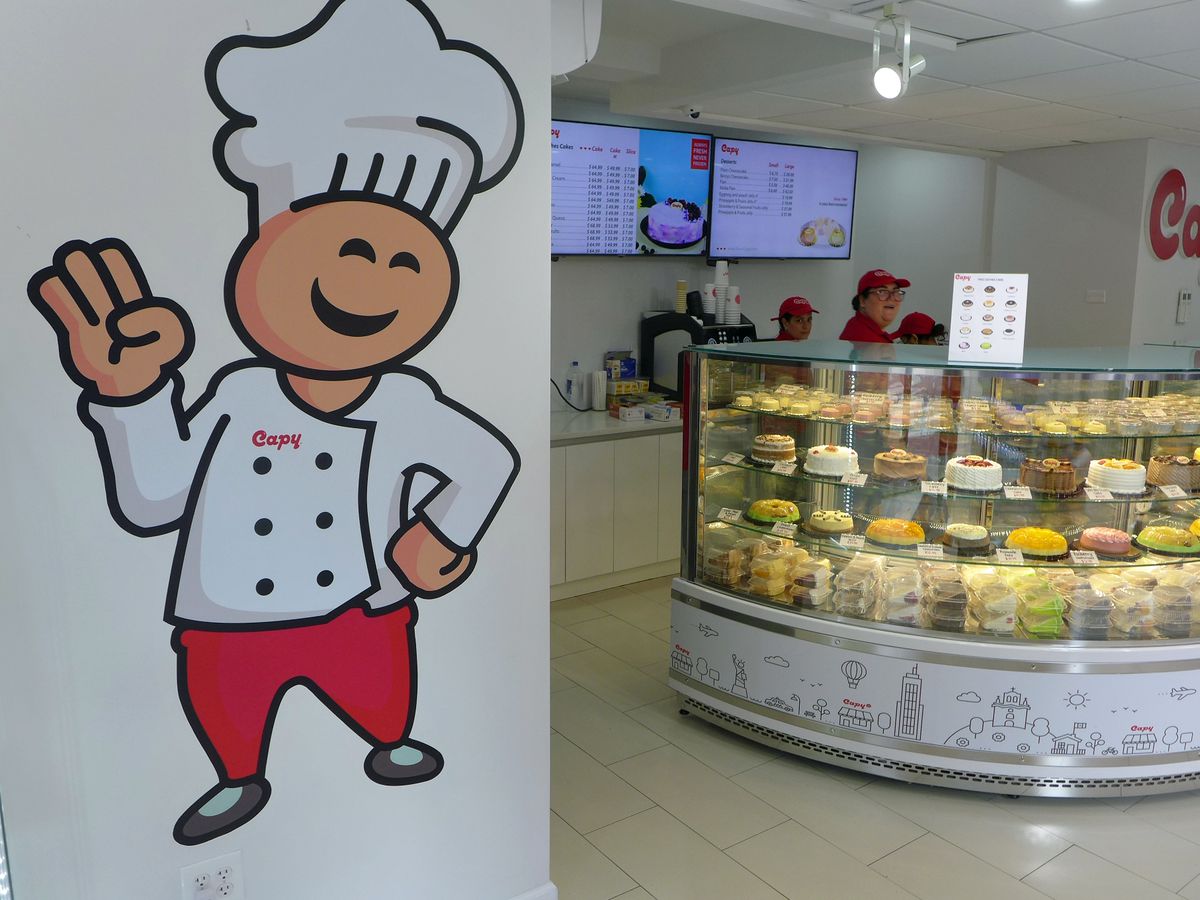 A cartoon figure in a chef’s hat at left, at right a curving case filled with spotlit cakes.