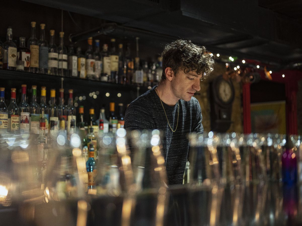 A bar worker stands behind a row of glasses. 