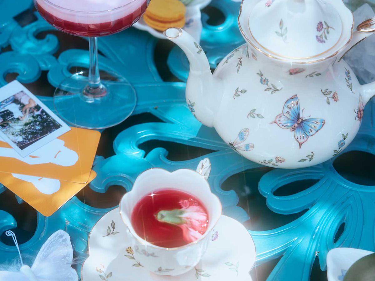 White tea pot with blue butterflies on a blue garden table next to a tea cup and cocktail glass filled with pink liquid with blue butterfly garnishes.