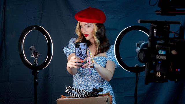 Danni Sanders (Zoey Deutch) posing in a red berett with a smart phone in front of two ring lights in Not Okay.