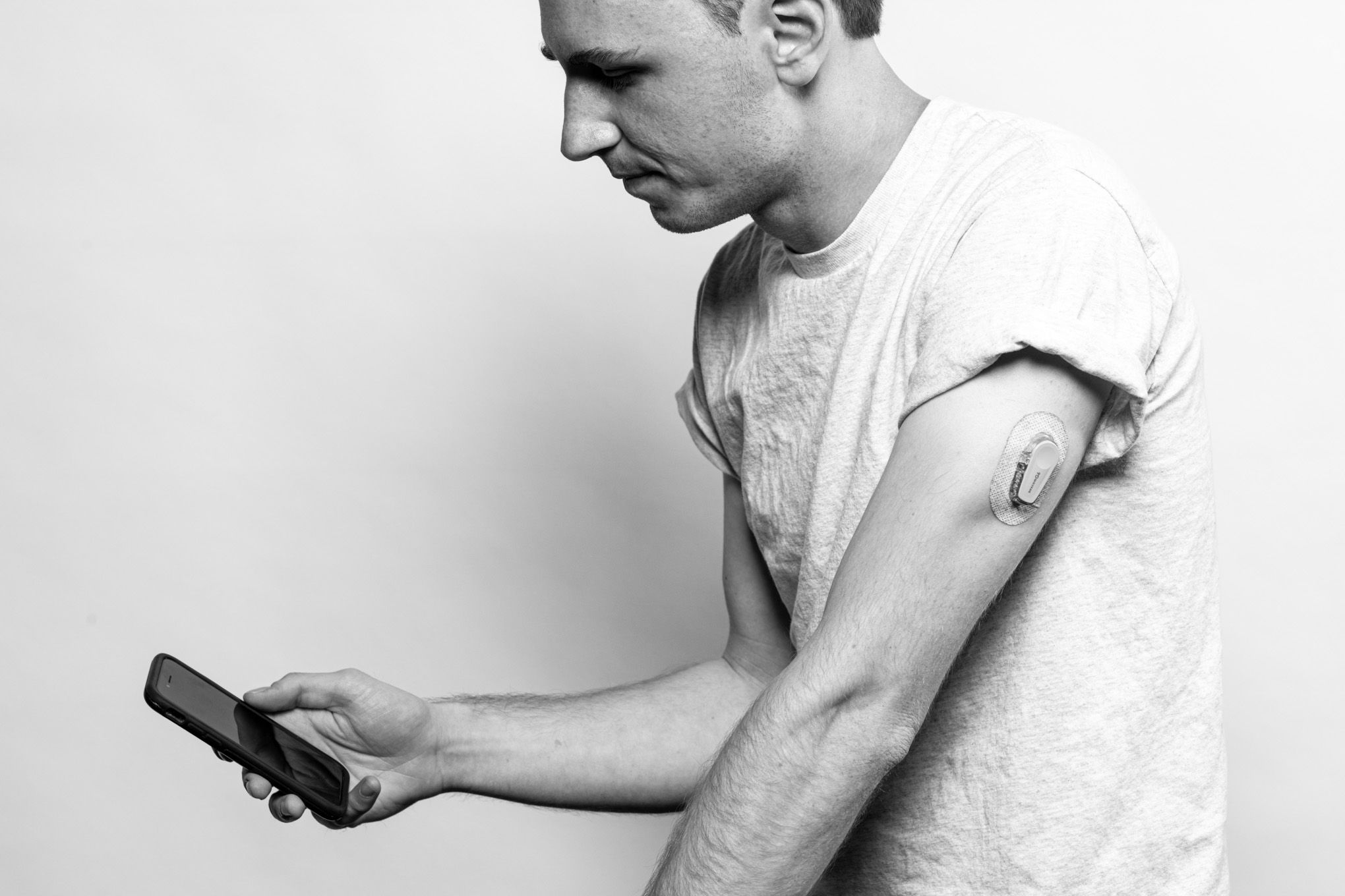 Une photographie en noir et blanc de John regardant son téléphone.  Il porte un glucomètre sur le haut de son bras gauche.