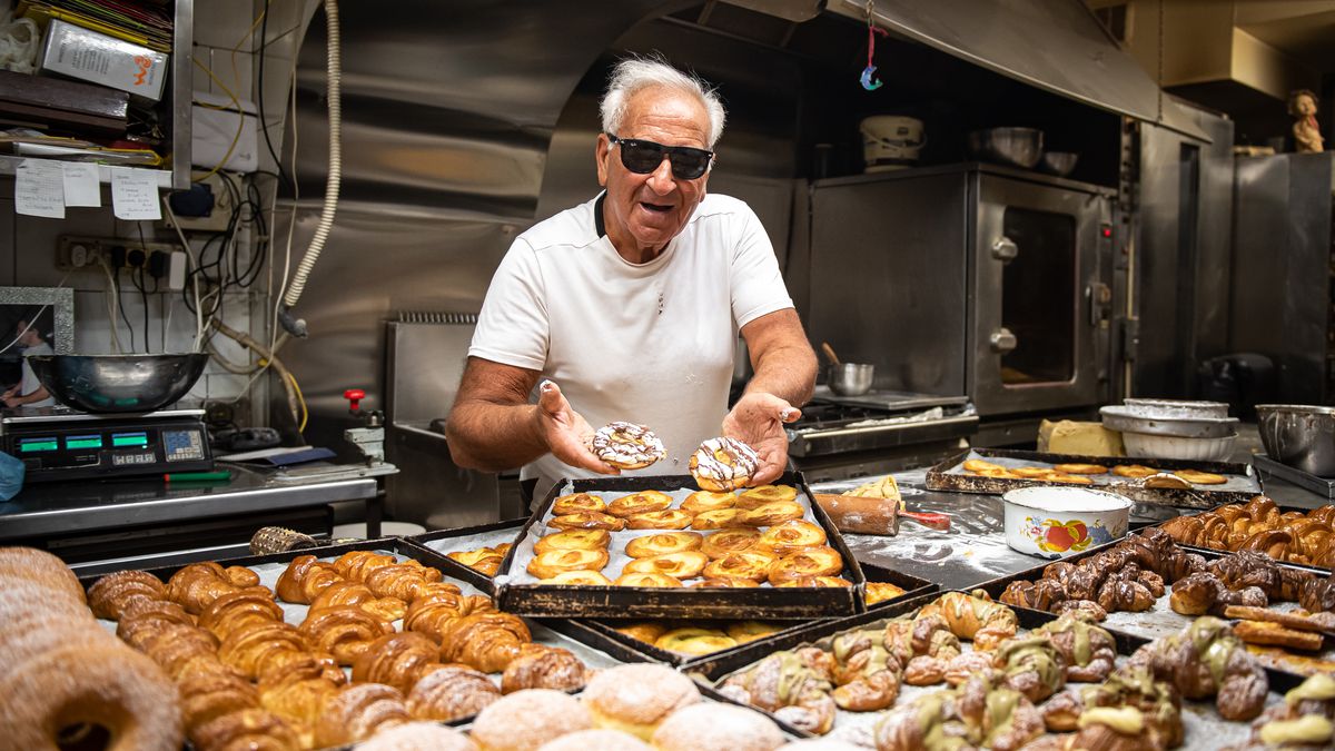 A baker in sunglasses holds up a few pastries from a massive table of assorted options in a bakery kitchen. 