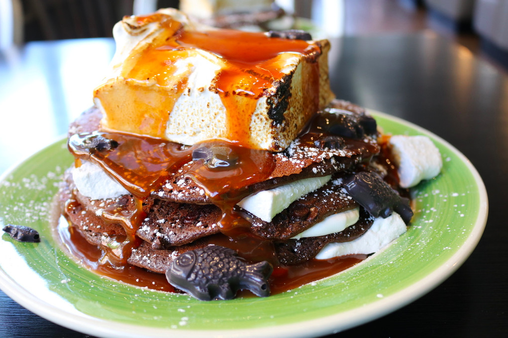 Stack of three pancakes with a variety of toppings and fillings, including caramel sauce, a giant block of toasted marshmallow, and chocolate fish