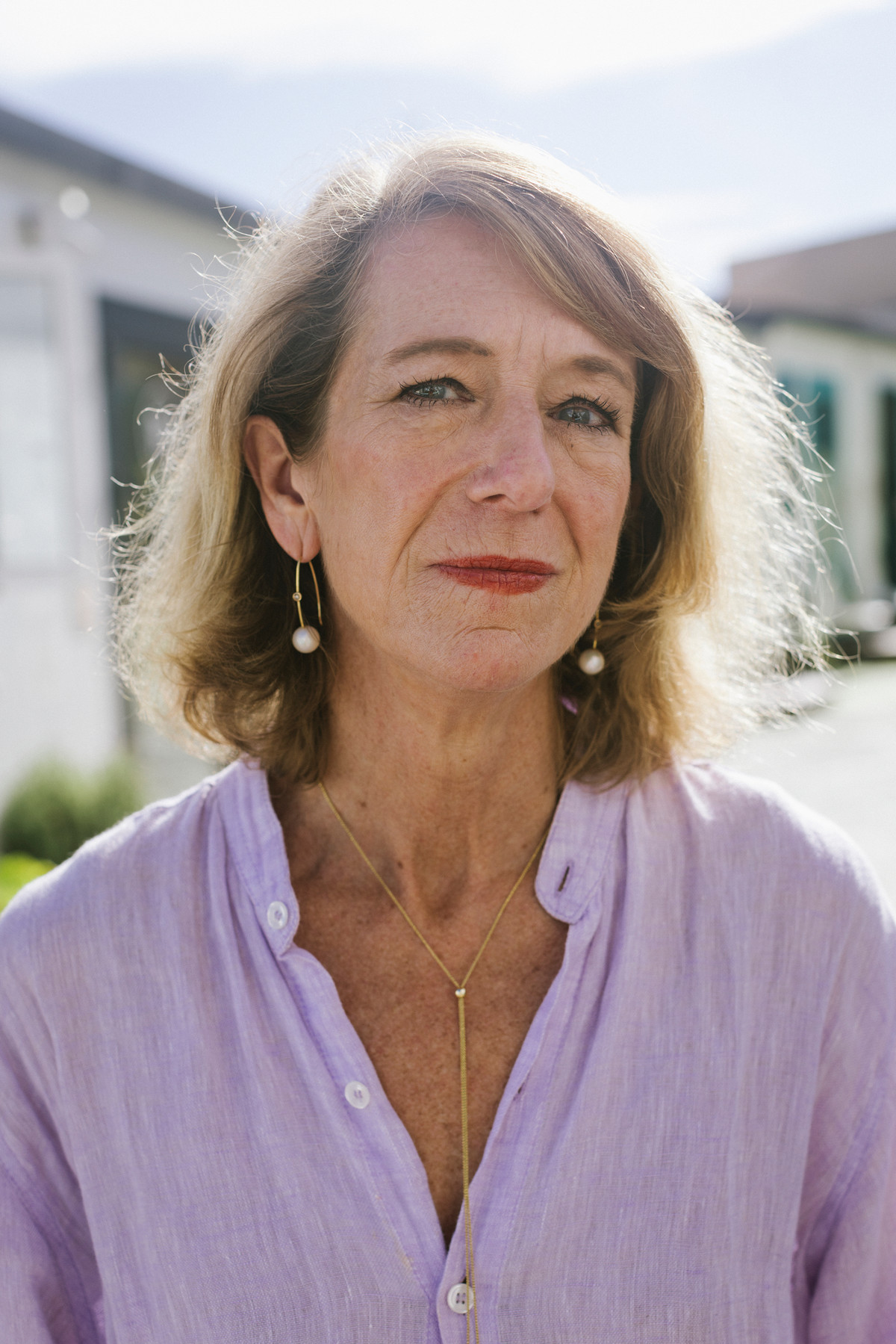 A middle-aged white woman with blond hair, wearing a lavender blouse and pearl earrings, looks into the camera.