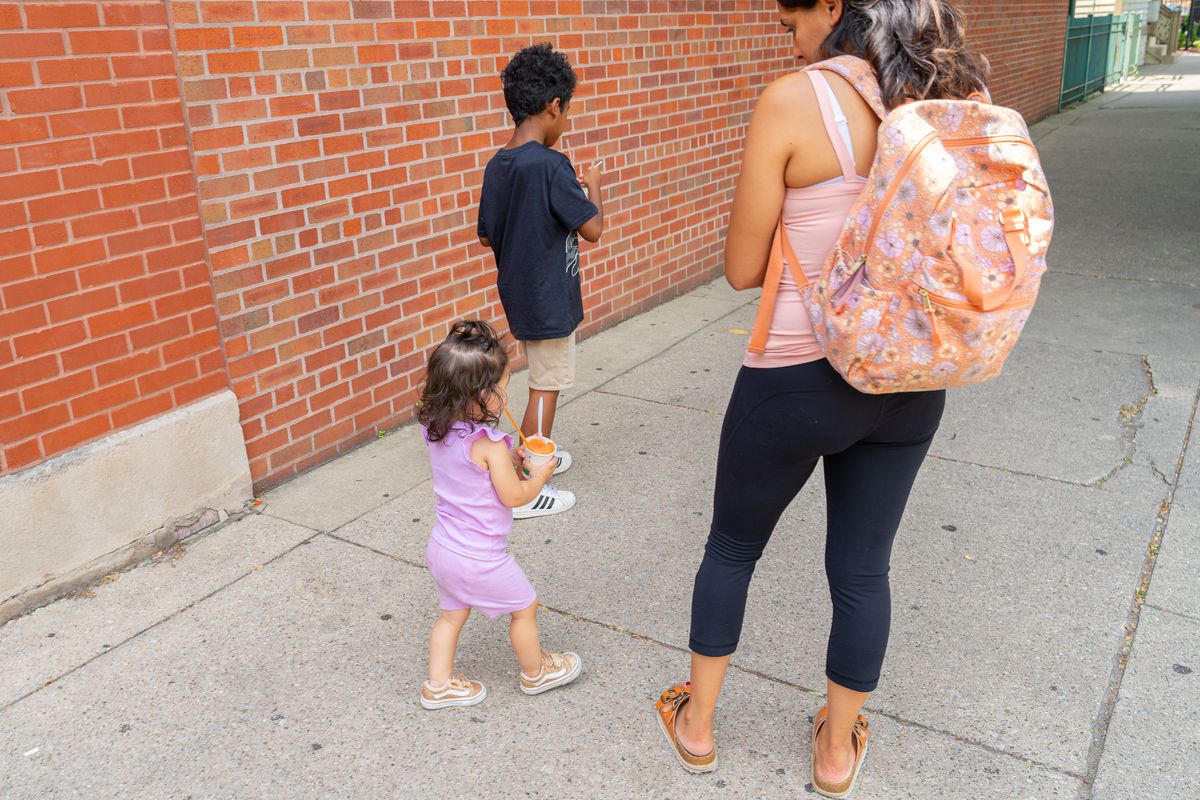 Two small kids holding cups of Italian ice walk down the sidewalk with an adult woman.