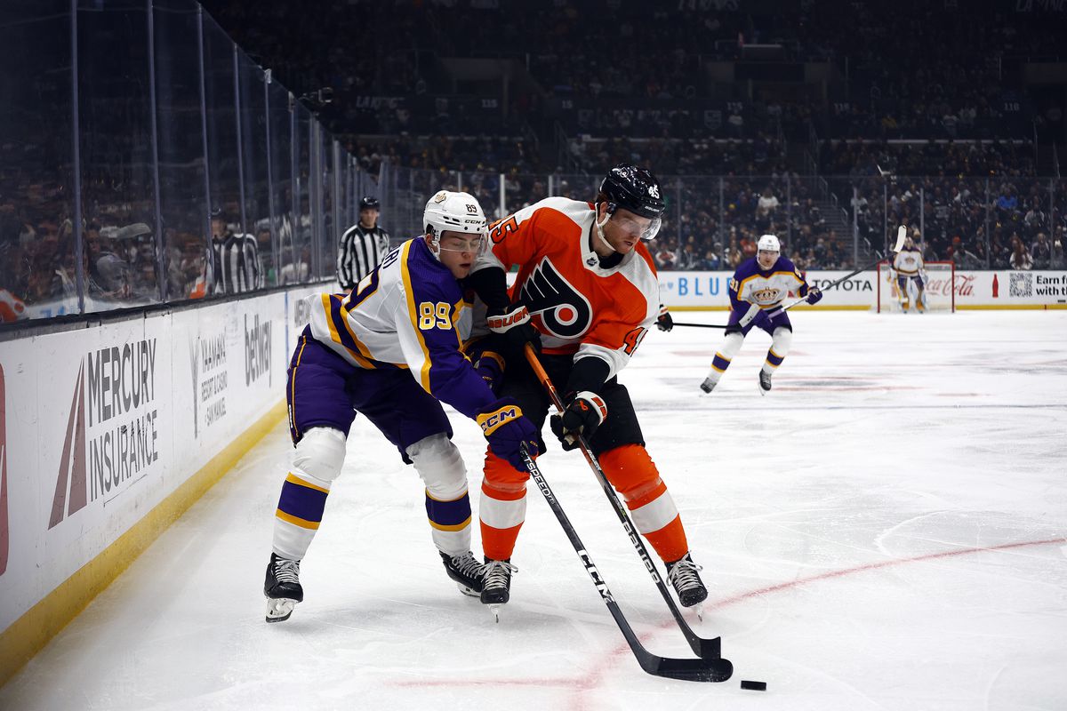 Noah Cates and Rasmus Kupair are pressed up into each other as they both battle for possession of the puck. Cates is in the Flyers’ orange jersey and Kupair is in the Kings’ white, purple, and yellow reverse retro jersey