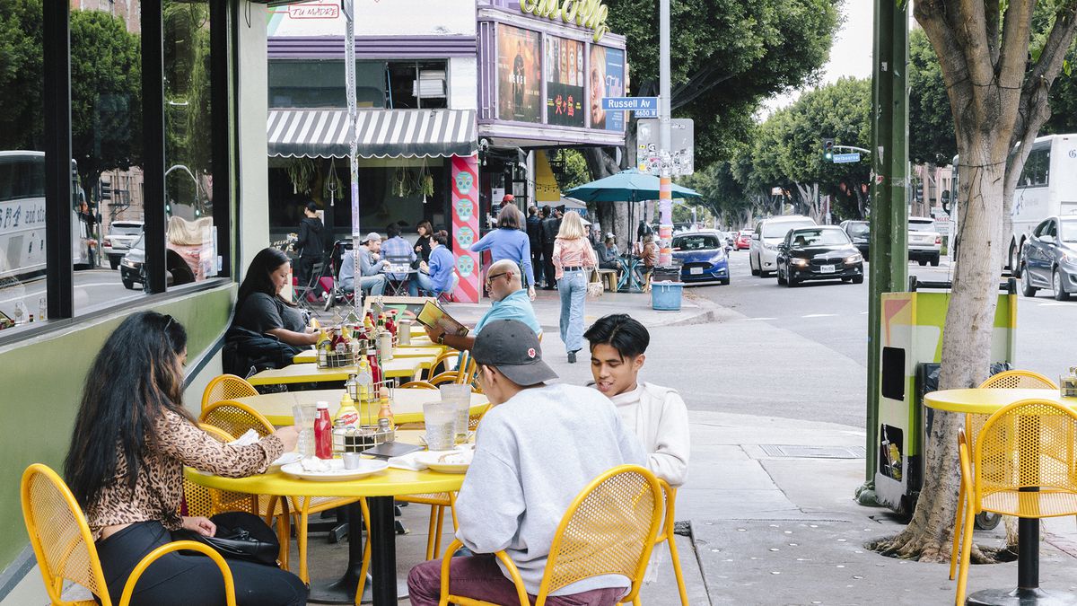 Young people sitting in yellow metal chairs dine on a sidewalk lined with trees. In the background, a vintage theater with green neon sign that reads in cursive print Los Feliz.
