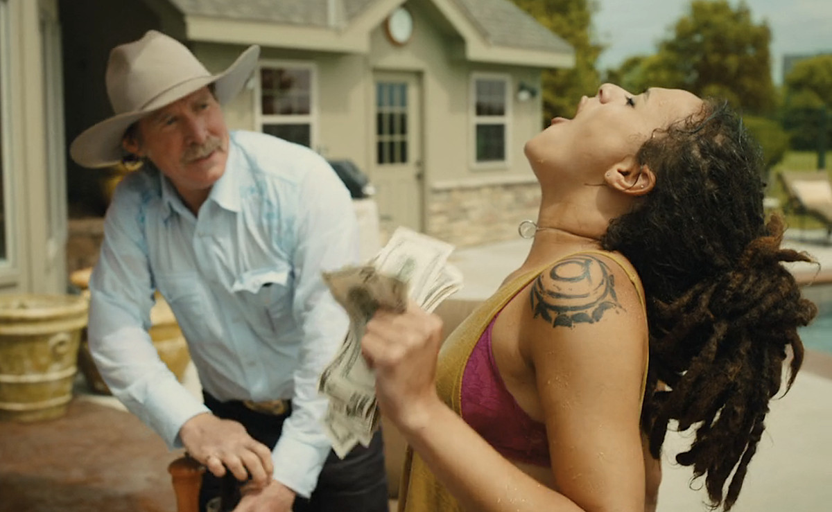 Star (Sasha Lane), a young woman in thick brown dreadlocks with a prominent black abstract circular tattoo on her shoulder, stands in front of a pool, grips a handful of $20 and $100 bills, throws her head back, and howls like a wolf in front of a fascinated-looking older white man in a white cowboy hat in a scene from American Honey