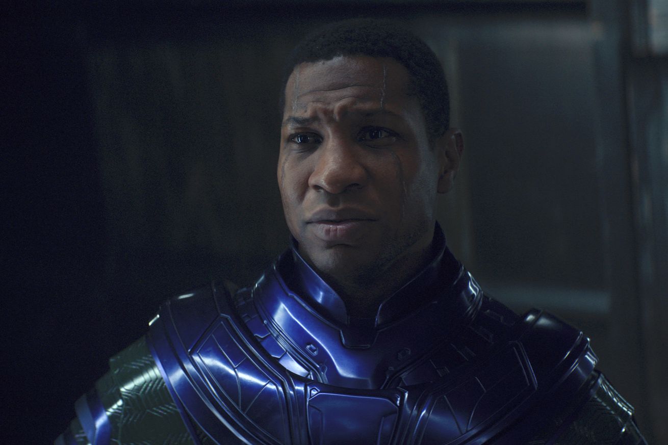 A man wearing gleaming armor that’s deep purple around his neck and shoulders, but deep emerald down his arms.