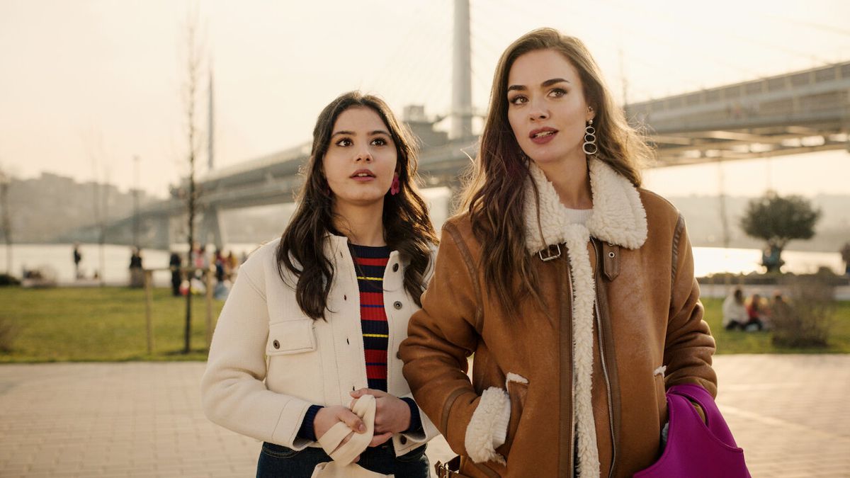 A young woman in a brown coat stands next to a younger girl in a white coat looking at something off-screen with a bridge visible in the background.