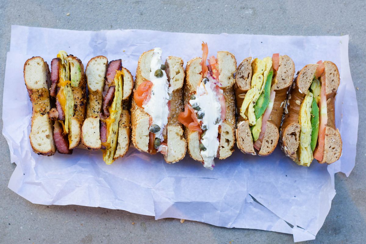 An overhead photograph of the cross sections of six bagel halves, filled with bacon, egg, cheese, avocado, tomato, and cream cheese
