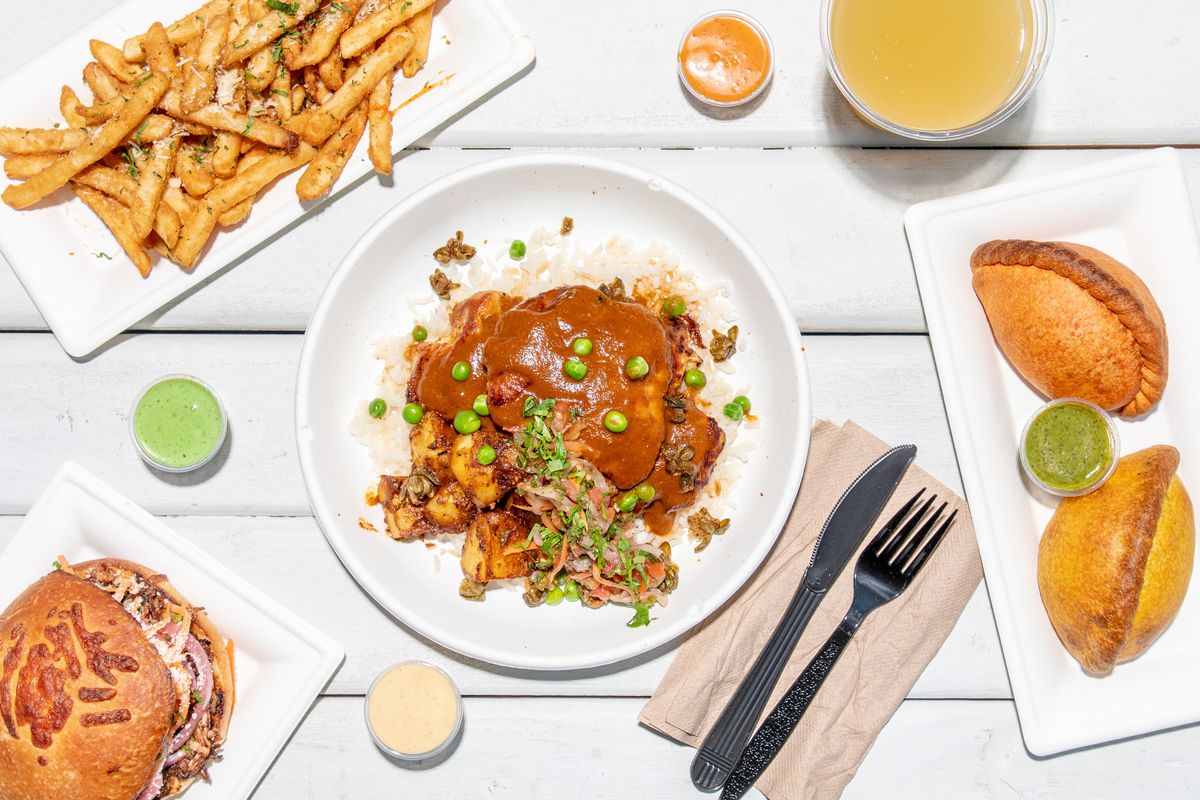 A spread of dishes included fries, a pork chola sandwich, golden saltenas, and a brown sauced picante de pollo in the center; all are photographed overhead atop white plates and a white table