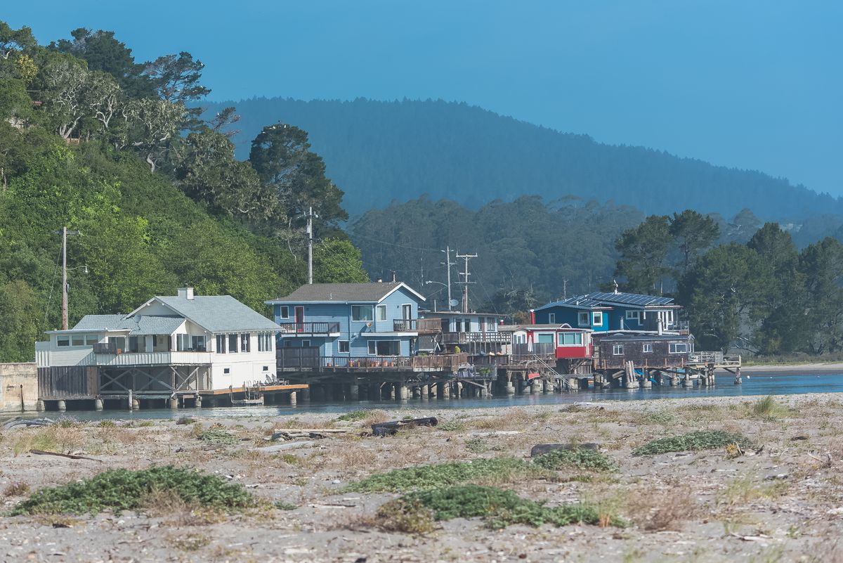 A handful of one- to two-story homes along the beach, with trees and mountainous terrain in the background.