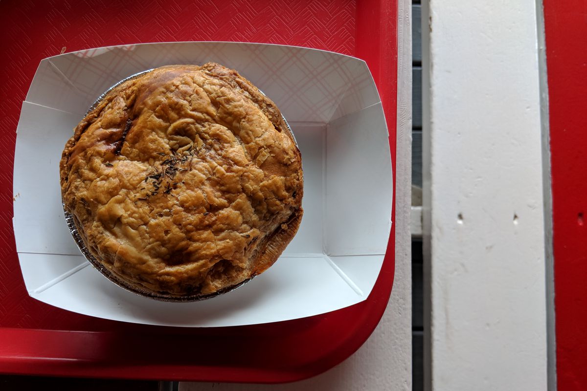 An Australian meat pie sits in a paper container on a red tray on a red and white picnic table