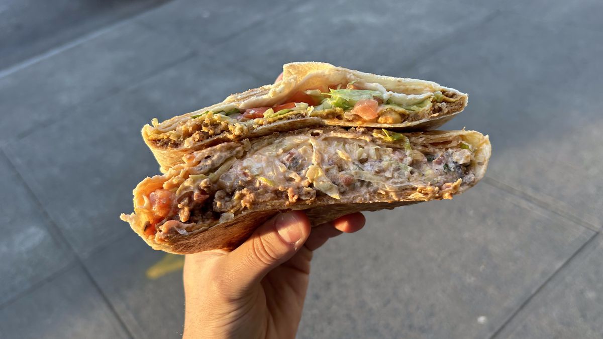 A hand clutches two Crunchwrap Supremes, which are cut in half to reveal greasy cross-sections.
