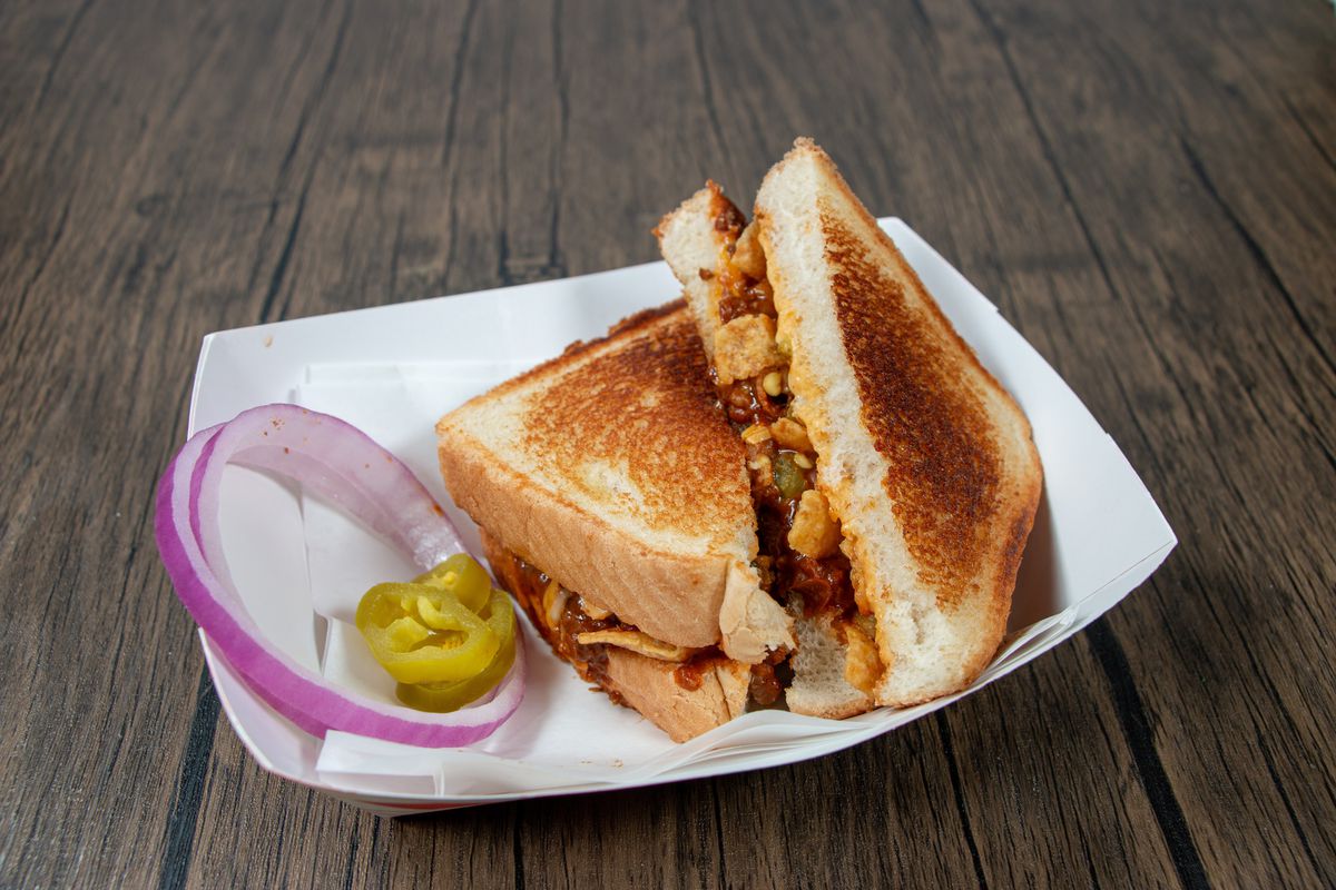 A grilled cheese sandwich with chili and Fritos layered inside sits in a red checked serving container with red onion slices and pickles on the side.