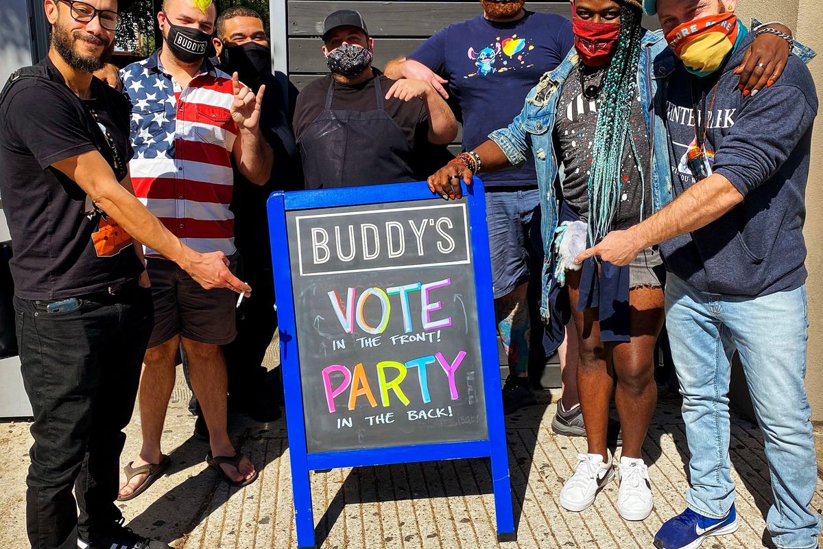 a group of masked people stand in front of a sign that reads “vote in the front, party in the back” outside a gay bar.