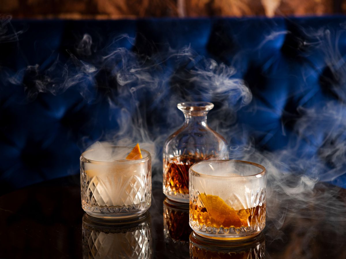 Three cocktails, some with smoke, on a dark wooden table next to a blue velvet booth.