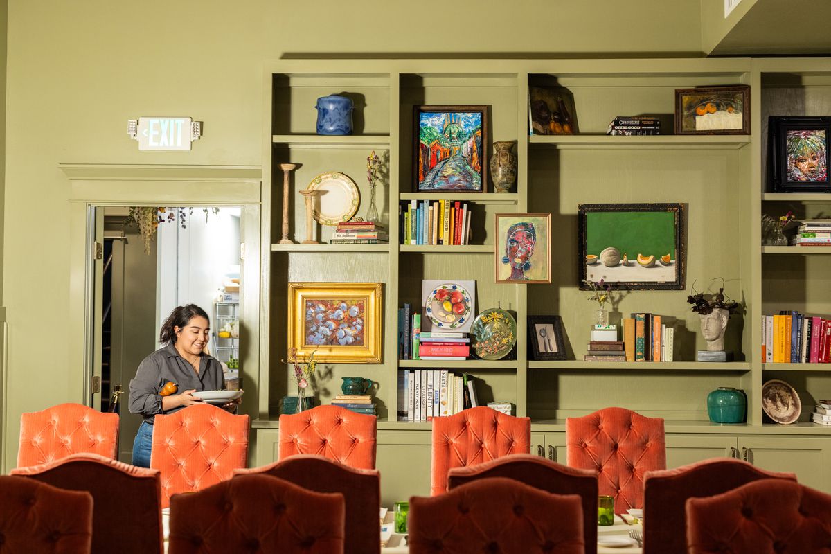 A server walks into a dining room with green walls and pink chairs. A large built-in book shelf loaded with books and art is visible in the background.