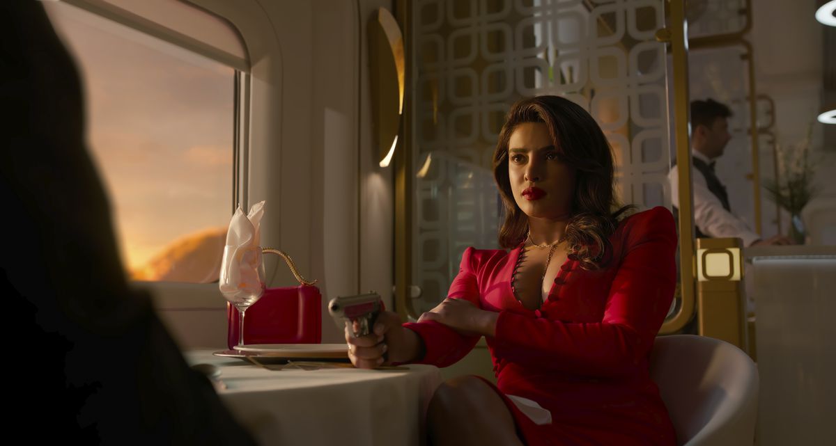 Priyanka Chopra Jonas as Nadia Sinh sits at a dining table on a train as the sun sets out the window, illuminating her red dress and the gun she’s pointing at a man off-screen across from her in the Prime Video show Citadel.