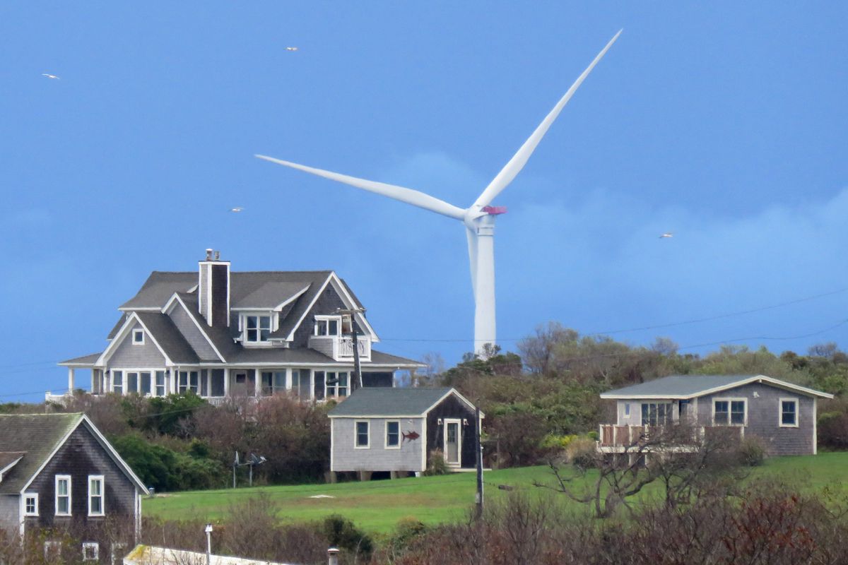 The large blades of a wind turbine rise over a hill with a few houses on it. The turbine dwarfs the houses, even though it is located in the water three miles away.