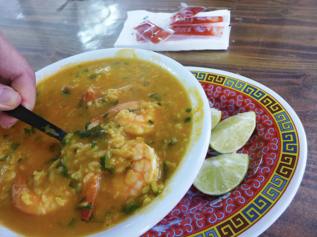 A rice-based yellow soup with hunks of shrimp is served in a bowl ontop of a red Chinese plate with three slices of lime.