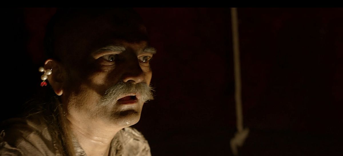 An old man with a white walrus mustache and a pearl earring stands in the dark, gaping at something offscreen, in the Indian horror film Tumbbad