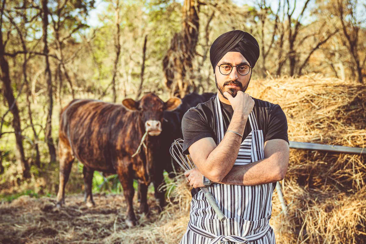chef Jassi Bindra stands in an apron outdoors in front of a cow.