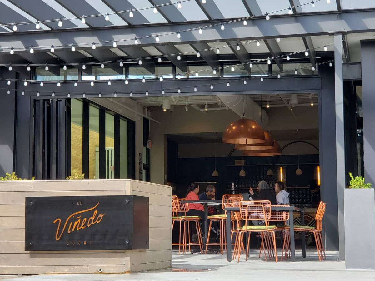 The patio at El Vinedo Local in Midtown Atlanta with orange metal chairs and wood tables. A dark metal sign etched with the El Vinedo Local wraps around the wooden wall entrance to the restaurant patio