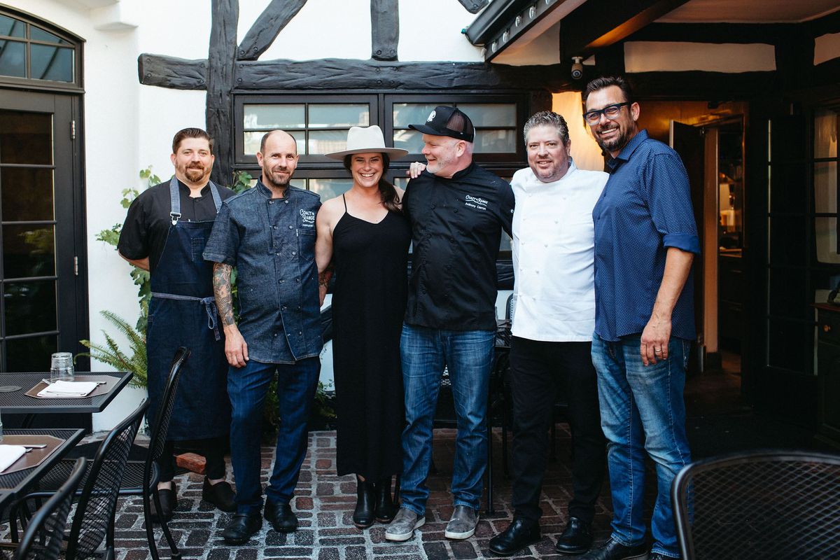 A group of chefs and owners stand holding sides dressed in mostly black on a patio.