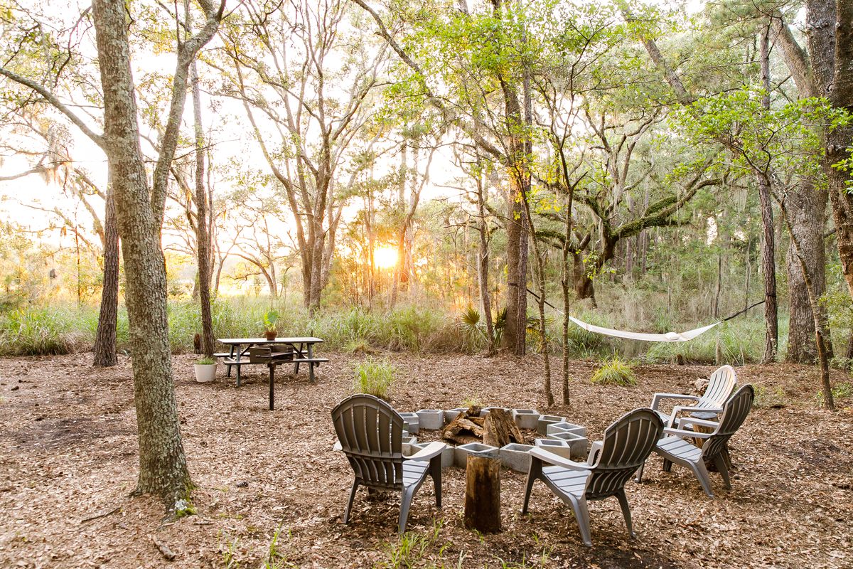 A cleared area in a forest with chairs facing a fire pit and a hammock sitting between trees in the distance as the sun sets.