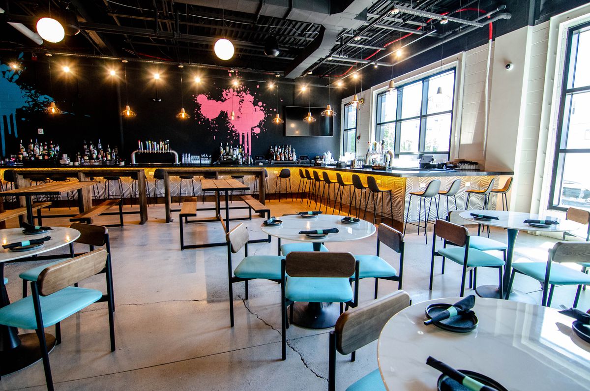 White marble four-tops with light blue chairs are in a dining area in front of a bar, which sits in front of a black wall with large blue and pink paint splatters.