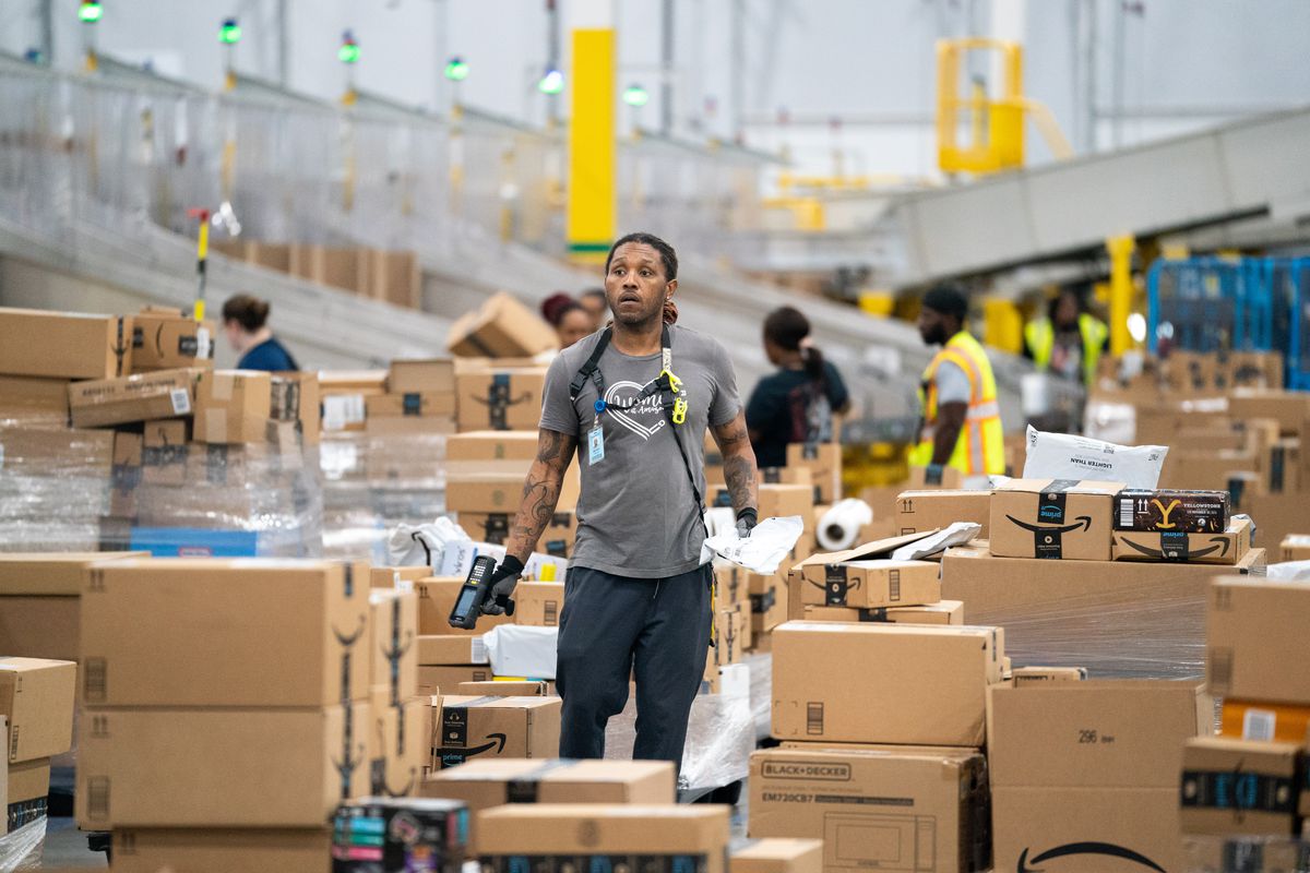 A worker stands amid many brown boxes in an Amazon facility.