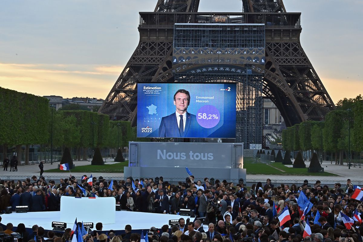 A large screen at the foot of the Eiffel Tower shows President Macron to a crowd of people.