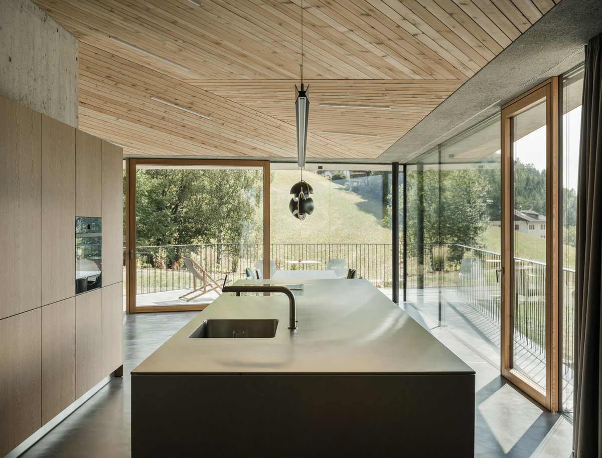 The kitchen area in a concrete home. There is an island with a sink, a wood ceiling and floor to ceiling windows overlooking a yard.