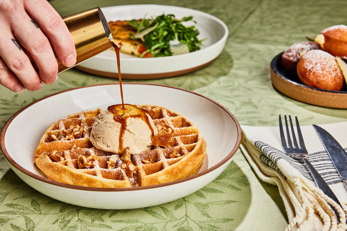 A dish with a waffle with ice cream in the center with espresso or syrup being poured over it. There’s also a salad and silverware on the table.