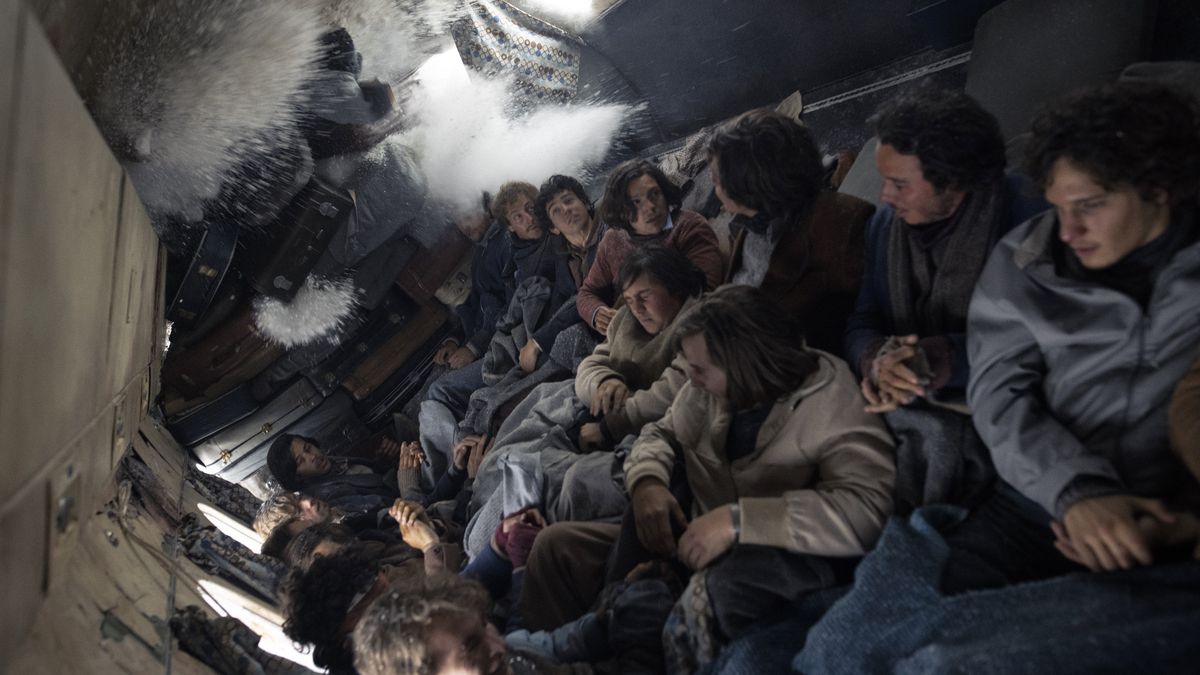 Fifteen rugby players in winter gear press together inside the shattered, overturned cabin of a plane as snow sprays through one end in the Netflix survival drama Society of the Snow