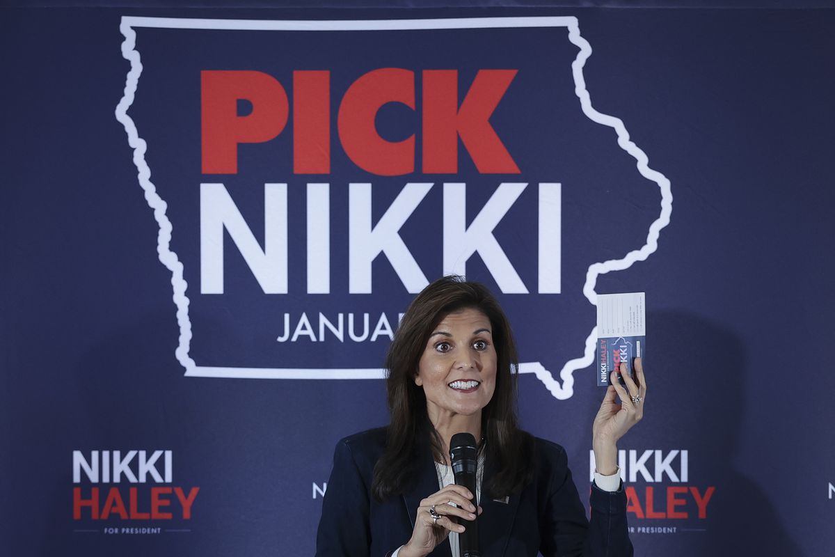 Haley smiles while holding a microphone and raising a card in her right hand. She stands in front of a navy blue backdrop that reads PICK NIKKI; the words are outlined by a silhouette of Iowa’s state boundaries.
