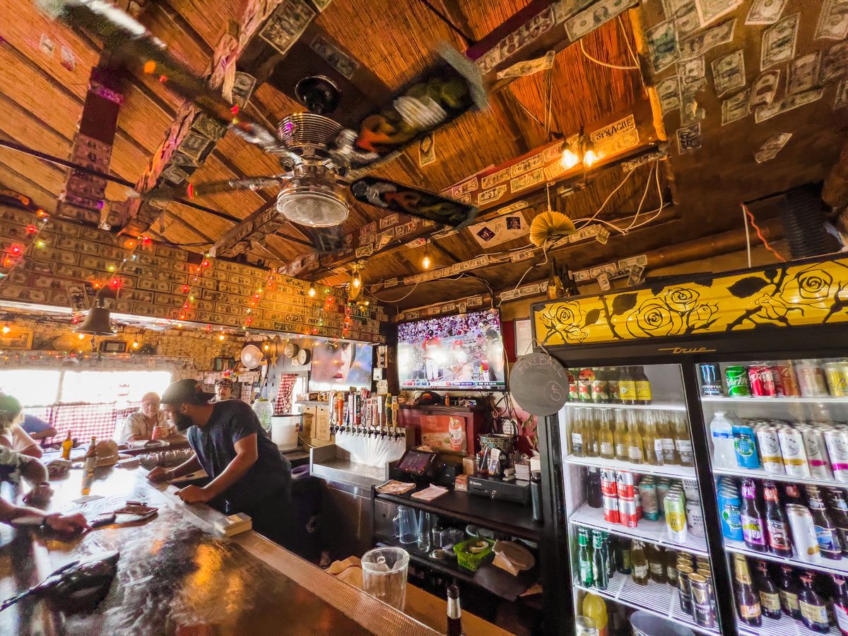 A bartender at daytime leans over a wooden bar at a dive bar to take a drink order.